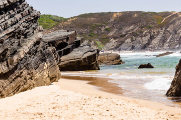 View of empty Praia da Amalia beach with ocean waves, cliffs and stones, wet golden sand and green vegetation at wild Rota Vicentina coast, Odemira, Portugal
