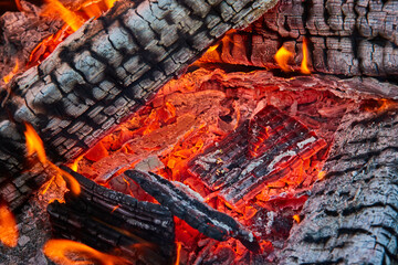 Close up of red embers beneath burning and charred black logs with white ashes and tiny flames