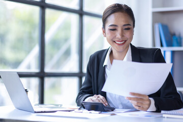 Young asian businesswoman sitting at desk with data and graph papers by using laptop at the office workplace, Accounting analyze, plan, and strategize about business improvement or finance. - Powered by Adobe