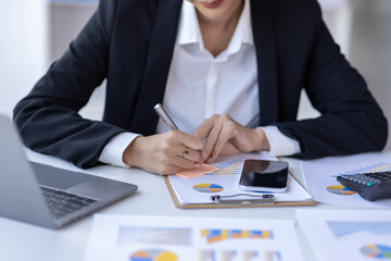 Young asian businesswoman sitting at desk with data and graph papers by using laptop at the office workplace, Accounting analyze, plan, and strategize about business improvement or finance.