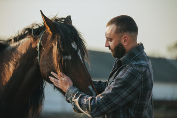 A man strokes a horse. A young bearded farmer takes care of horses.
