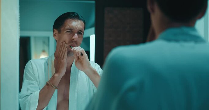 Dental problems and toothache. Reflection in the mirror millennial male in bathrobe brushing teeth with toothbrush in bathroom and stops because toothache. Male suffering from teeth pain.