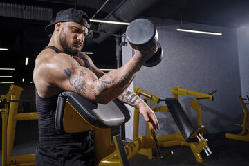 Bearded athlete in black vest and cap. He is lifting a dumbbell, training his biceps, sitting on...