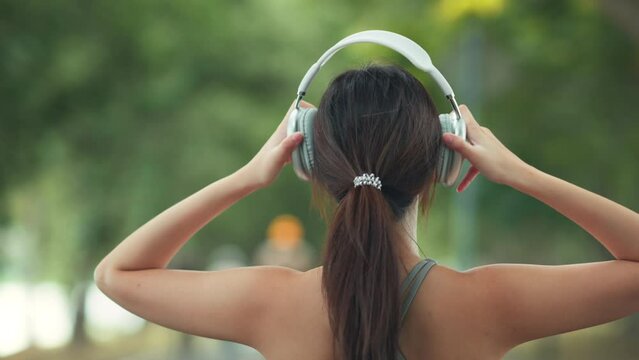 Happy asian woman adjusting wireless headphones before starting jogging and listening to music on running route surrounded by nature. Slow motion.