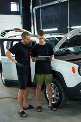 Two auto mechanics stand next to a white SUV with the hood open and look at the tablet they hold in their hands while repairing the car