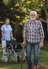 Fototapeta na wymiar Senior man walking with stick in front of wheelchair
