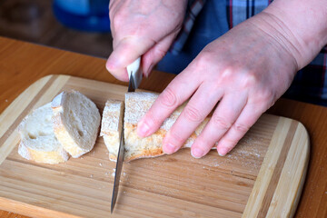 The hands of an elderly woman cut white wheat bread baguette