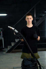 Portrait of young car wash worker holding a vacuum cleaner and showing a thumbs up sign on the...