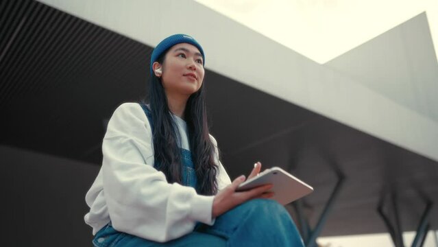 Cute Asian young woman in a hat with a tablet on the street. Drawing on a graphics tablet. Freelancing and earning online.