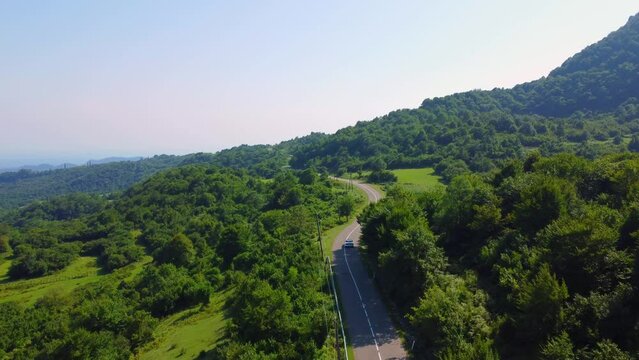 Aerial view of convertible car driving along country road in forest in mountains of Georgia in summer on sunny day. Cinematic drone captures shot of beautiful empty road. Camera flies behind car