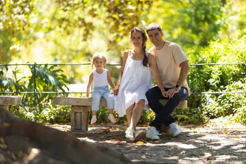Fototapeta premium Family in park - a little girl with her parents sitting on a bench and looking in the camera