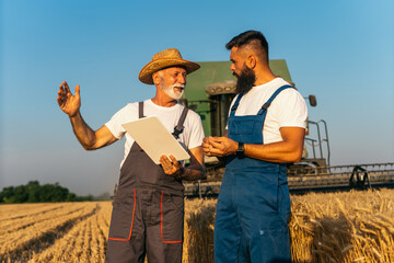 Naklejka na ściany i meble Grandfather and a grandson standing in wheat field and conversation about harvest. They using digital tablet. Combine harvester working in background. Family business concept.