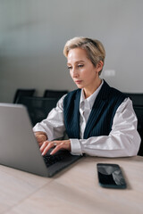 Vertical shot of confident middle-aged 50s businesswoman in suit sitting at modern office desk working on laptop do freelance online job. Senior adult business woman work on project typing on computer