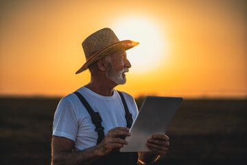 An elderly man stands in a field being plowed, holding a digital tablet that helps him monitor farm...