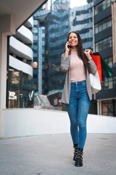 Full Length Photo Of Female Figure Walking Down The Street In Urban City Area. Modern Woman Having A Phone Call Outdoors While Holding Gift Bags.