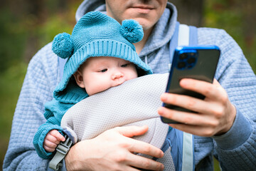 Father and newborn baby in a sling in the autumn forest, man with mobile phone. Happy family.