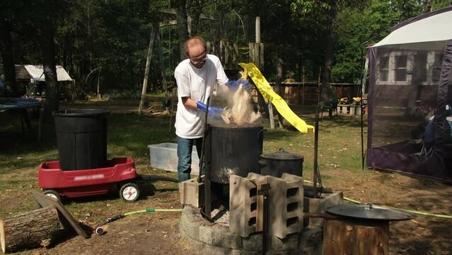 Man scalding butchered chicken using large pot with heated water over fire pit for easy feather plucking. Processing poultry traditional way by hand.