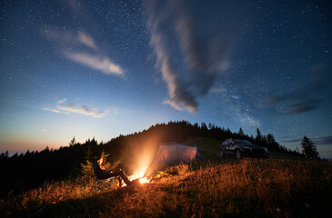 Camping in mountains under evening cloudy sky. Young man having a rest next to campfire, near tourist tent and off-road car on hill. Travelling man sitting and stargazing at night. - obrazy, fototapety, plakaty