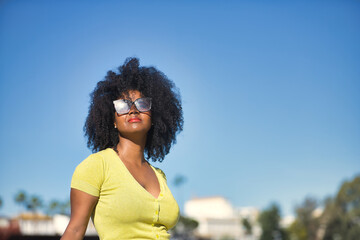 Portrait of young, beautiful, black woman with afro hair, with yellow t-shirt and sunglasses, looking at the sun, relaxed and calm. Concept vacation, travel, sun, sunglasses, current, modern.