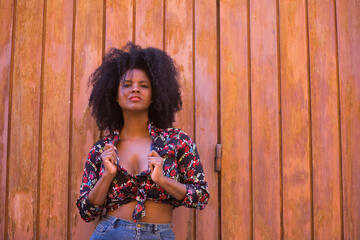 Fototapeta premium Young woman, beautiful and black with afro hair, with a floral shirt leaning on a wooden board door, in a sensual, provocative and flirtatious attitude. Concept beauty, sensuality, provoke, flirt.