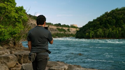 Man standing near a river, capturing photographs with a digital camera