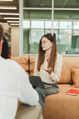 Serious pensive young woman using laptop sitting on sofa in modern office explaining something