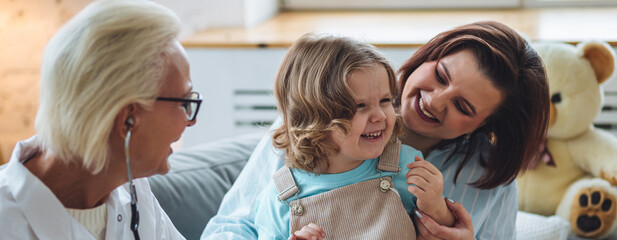 Concept healthcare medical assistance, insurance. Kind senior female paediatrician doctor examining little child sitting on mother's laps, during home visit or clinic check up. Banner