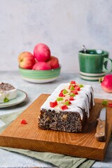 Polish poppy seed cake covered with white sugar glaze and decorated with candied fruits, on a wooden board on a gray concrete background. Poppy baked goods.