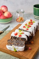 Polish poppy seed cake covered with white sugar glaze and decorated with candied fruits, on a wooden board on a gray concrete background. Poppy baked goods.