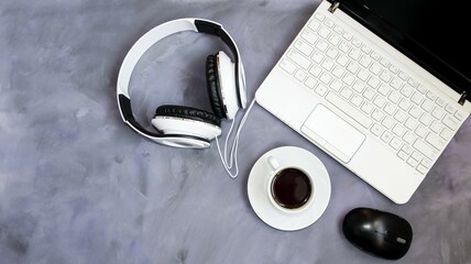 On a table on a gray background is a laptop and a white cup of coffee and a computer mouse, headphones, coffee break concept, office work, online learning, webinars.  Top view, flat lay.
