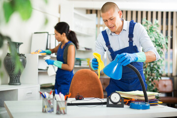 Skillful worker of cleaning service wearing uniform and rubber gloves wiping dust on furniture with disinfectant in office