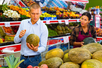 Asian woman in uniform helping client to choose ripe melon in supermarket.