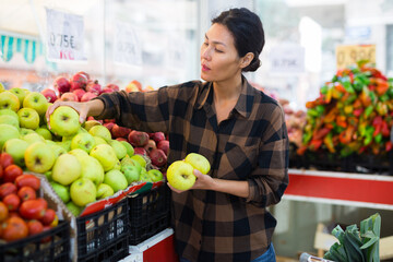 Portrait of attractive adult asian woman choosing sweet apples on counter of farmers market..
