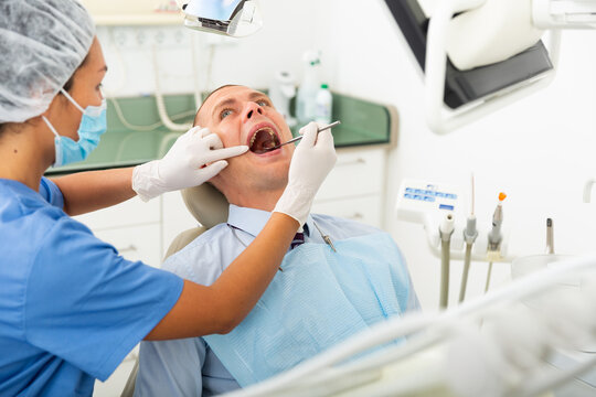 Woman dentist in a protective mask working at the clinic conducts an examines a man patient with the help of tools sitting ..in a dental chair. Close-up image
