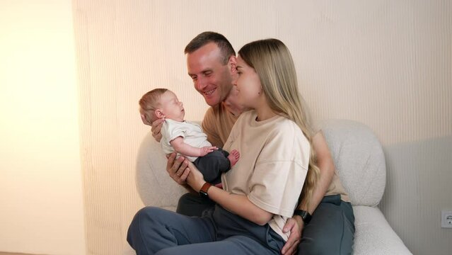Beautiful Caucasian couple sitting on the sofa with their newborn. Happy dad kisses the sleeping infant son.