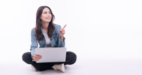 Cheerful asian woman using laptop pointing finger looking empty copy space sitting on floor over isolated white background. Positive student teenager use finger pointing present product with computer.