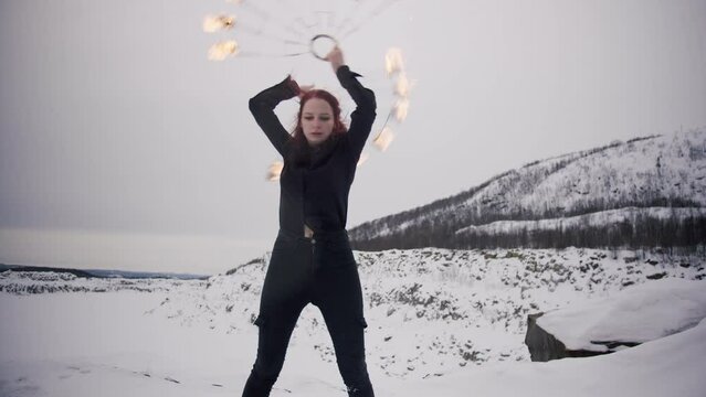 A young caucasian girl with red hair in a black shirt and black jeans masterly spins staff fire fans in the winter forest with snow and hills. Perfomance with staff in a fire show