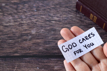 God cares for you, handwritten text note on human hand and holy bible book on wooden table. Top...