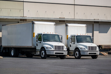 Day cab middle duty rig semi trucks with long box trailers loading cargo standing side by side in...