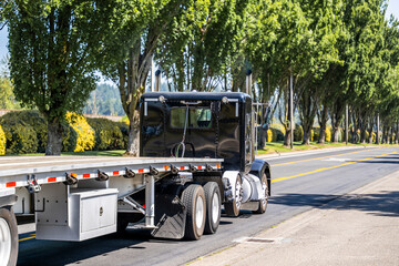 Low cab profile black classic American big rig semi truck transporting empty flat bed semi trailer...