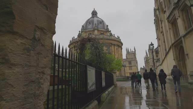 People students pass Radcliffe camera library building dome and exterior metal fence. Study in Oxford university and british education concept