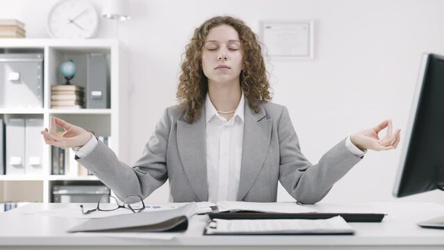 Young businesswoman wearing white shirt and jacket practice yoga and deep breathing exercises at office desk to combat fatigue