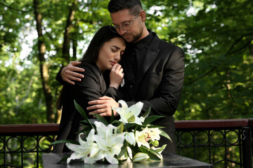 Sad couple mourning near granite tombstone with white lilies at cemetery. Funeral ceremony
