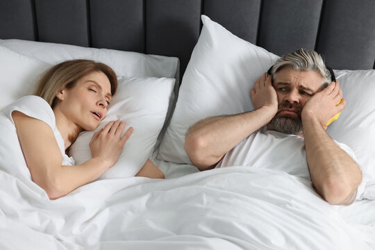 Irritated man with headphones lying near his snoring wife in bed at home