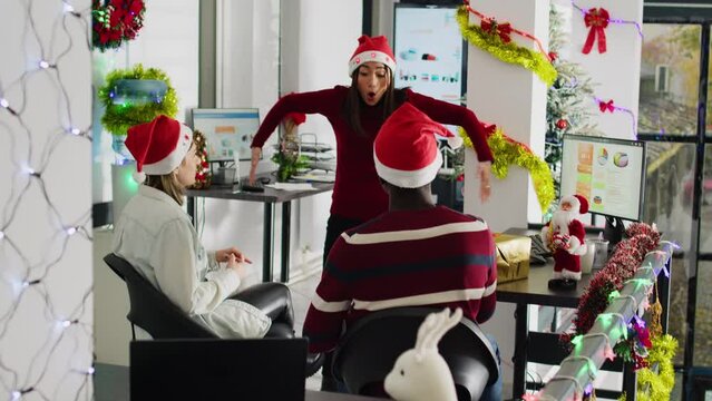 Diverse workers wearing Santa hat enjoying Christmas spirit mood playing pretending acting game during break. Coworkers having fun doing funny gesticulations during pantomime in xmas adorn office