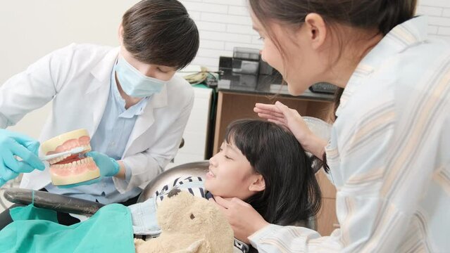 Asian male pediatric dentist demonstrates toothbrushes to a girl from teeth implant model with her mother in dental clinic for hygiene. Professional orthodontic oral healthcare work in a kid hospital.