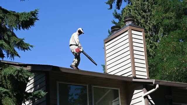 Senior man with a gas powered leaf blower cleaning out debris in a roof gutter on a residential building
