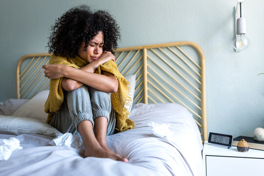 Young multiracial woman feeling sick and cold sitting on bed covered with blanket, choking temperature with thermometer.