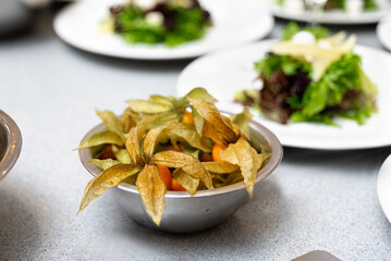 a close up of a bowl of unpeeled physalis berries on a table, big leaves foliage and stems on a table in a kitchen