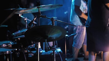 Close-up of drum kit cymbals shrouded in smoke. Band performance in the background. Drumsticks sometimes hit the cymbal. The stage is illuminated by multi-coloured lights. Live performance